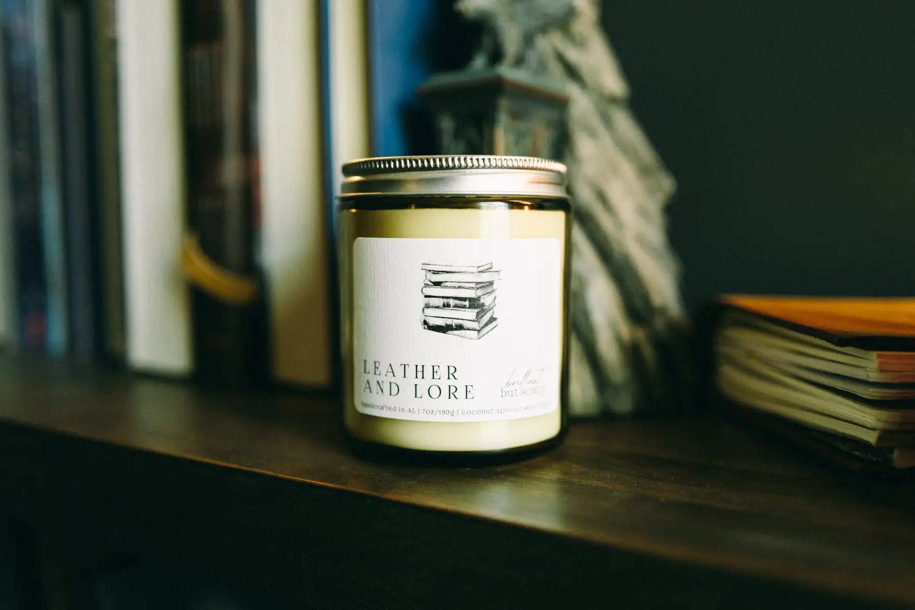 Leather and Lore fantasy candle in oatmeal jar on dark wood bookshelf with books - Brilliant but Scary
