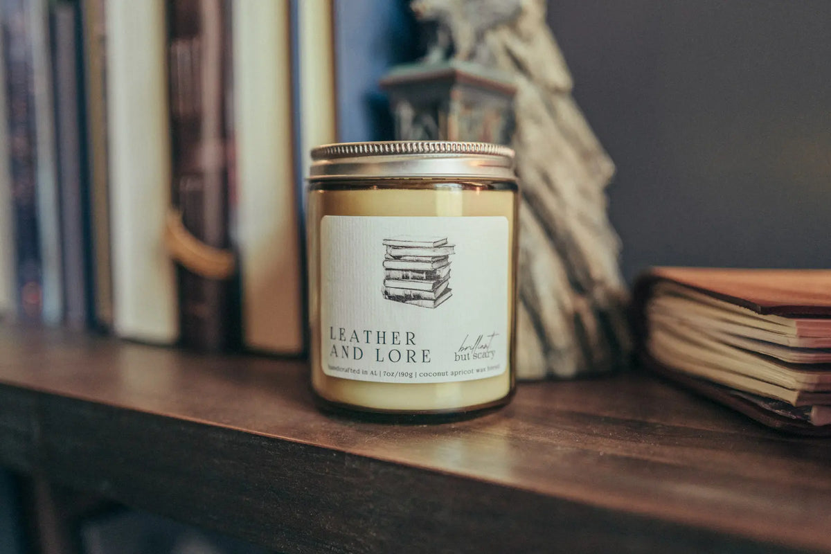 Leather and Lore fantasy candle in oatmeal jar on dark wood bookshelf with books - Brilliant but Scary