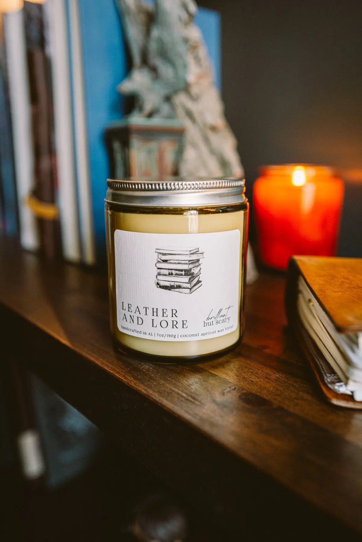 Leather and Lore fantasy candle in oatmeal jar on dark wood bookshelf with books - Brilliant but Scary
