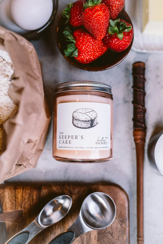 The Keeper's Cake fantasy candle in pink jar on counter with a wand and baking ingredients - Brilliant but Scary